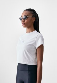 White cropped T-shirt with a crew neckline, featuring a small blue logo. Worn with high-waisted black leggings and oval sunglasses.