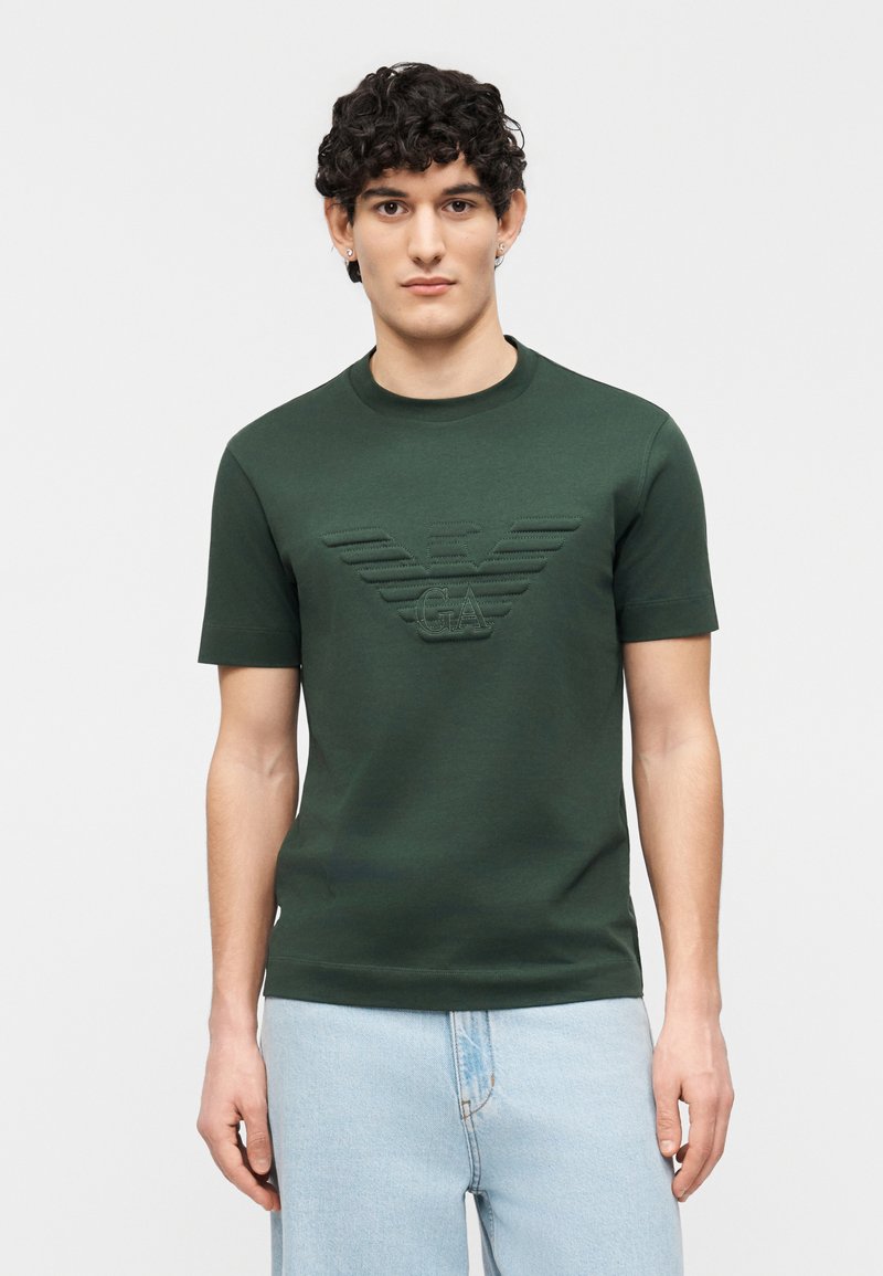 Νεαρός άνδρας με σγουρά σκούρα μαλλιά φοράει σκούρο πράσινο κοντομάνικο μπλουζάκι με ανάγλυφο μεγάλο λογότυπο με φτερά και ανοιχτόχρωμο μπλε τζιν, μπροστά σε λευκό φόντο.