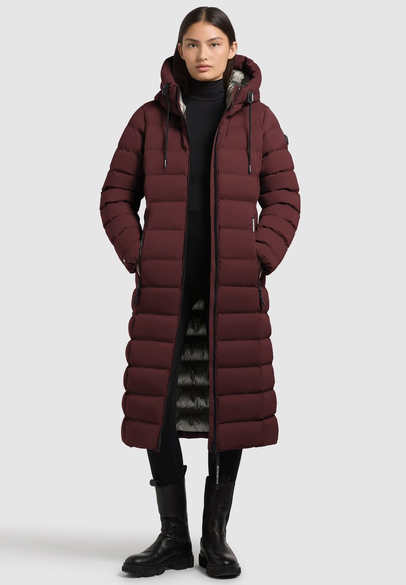 Giacca a vento maroon con un design lungo, cappuccio e dettagli con zip. Presenta imbottitura trapuntata e una fodera interna a contrasto. Abbinata a leggings neri e stivali.