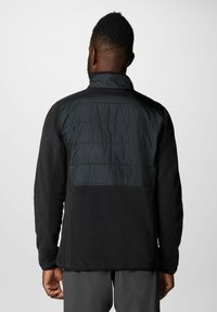 Giacca nera in tessuto texturizzato, con una parte superiore liscia e imbottita. Il colletto è dritto, con maniche lunghe e un taglio dritto.