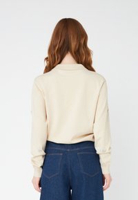 Maglione beige in maglia con colletto, maniche lunghe con dettagli decorativi ai polsini, abbinato a pantaloni a gamba larga blu scuro.