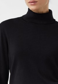 Nahaufnahme einer Person, die einen schwarzen Rollkragenpullover trägt und Hals sowie Oberkörper vor einem einfarbigen Hintergrund zeigt.