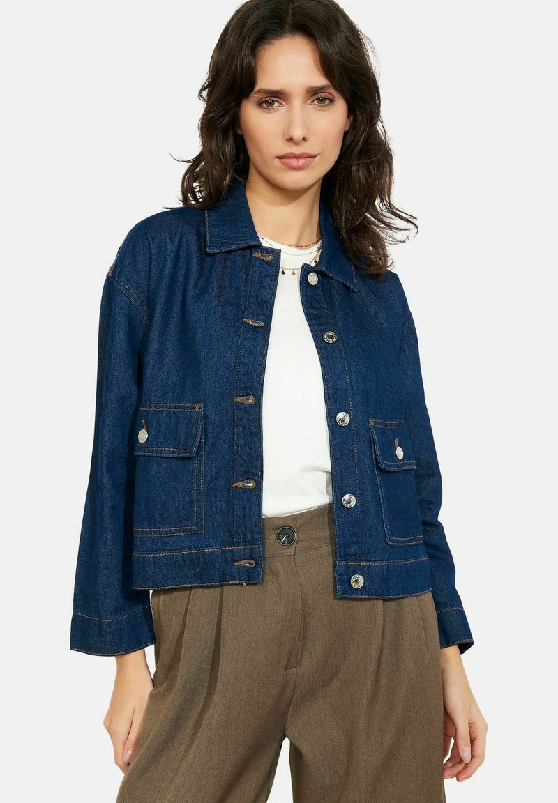 Žena v tmavě modré džínové bundě se stříbrnými knoflíky přes bílý top, kombinovaná s hnědými kalhotami s vysokým pasem, stojící před jednoduchým pozadím.