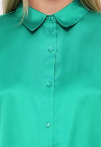 Camisa de colarinho turquesa com um tecido suave e brilhante. Frente com botões e botões combinantes, além de um colarinho estruturado.