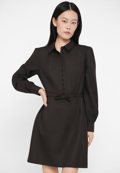 Sieviete ar taisniem melniem matiem, ģērbusies melnā garās piedurkņu kleitā ar auduma pogām un sasietu jostu, nostājusies pozā ar vienu roku uz gurniem.