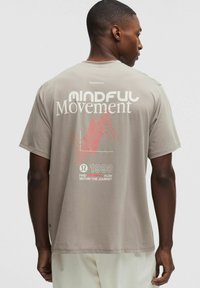 Mężczyzna w beżowej koszulce lululemon z napisem "Mindful Movement" i czerwonym wzorem fali na plecach, noszący jasne spodnie.