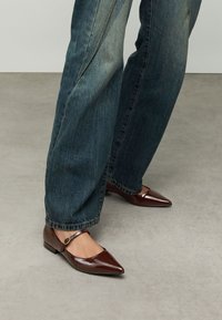 Bruine schoenen van lakleer met puntige neuzen en een dunne enkelband met een ronde gesp, gecombineerd met blauwe spijkerbroeken.