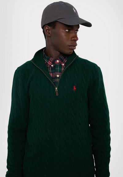 Green cable-knit sweater with a zip collar, featuring a red logo. Underneath, a plaid button-up shirt with red and green tones. Gray cap on head.