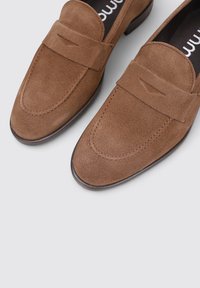 Mocassins en daim marron avec un léger talon, présentant un embout arrondi élégant et une bande décorative sur le devant. Doublure intérieure lisse.