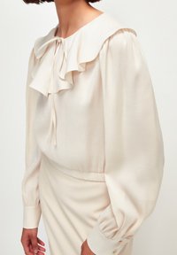 Blusa color crema con colletto arricciato, maniche lunghe a sbuffo e un laccio al collo. Realizzata in un tessuto leggero e morbido.