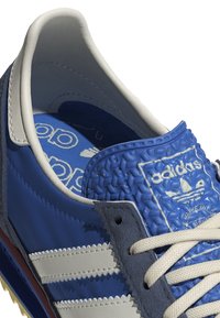Gros plan sur une basket Adidas bleue avec languette bleue texturée, lacets blancs, bandes blanches sur le côté et logo Adidas visible sur la semelle intérieure.