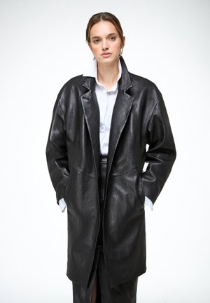 Jonge vrouw met een oversized zwarte leren jas en een wit overhemd met kraag, staand met handen in de zakken tegen een effen achtergrond.