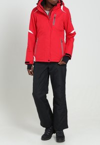 Rote isolierte Jacke mit Kapuze, ausgestattet mit weißen Akzenten und mehreren Reißverschluss-Taschen. Getragen mit schwarzen Hosen und schwarzen Stiefeln.