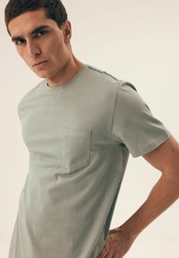 Jasnozielona bawełniana koszulka z okrągłym dekoltem, krótkimi rękawami i kieszonką na piersi, z subtelnymi detalami ściegów na ramionach.