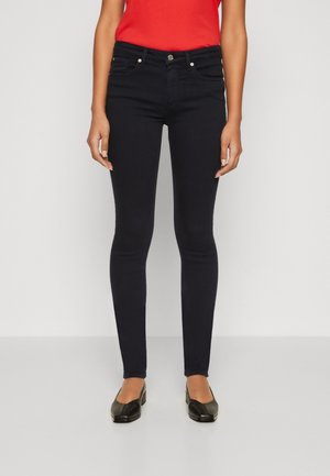 Femme portant un jean skinny noir, un haut rouge et des chaussures noires à petit talon, debout devant un fond clair uni.