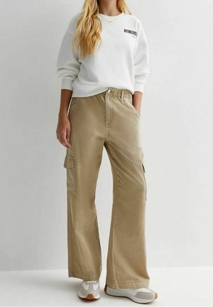 Vrouw met een witte sweatshirt met lange mouwen en beige wijde cargobroeken met zakken, staand tegen een effen achtergrond.