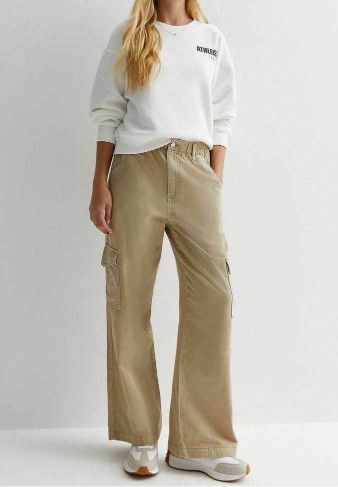 Femme portant un sweat-shirt blanc à manches longues et un pantalon cargo beige à jambes larges avec poches, debout devant un fond uni.