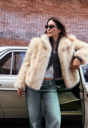 Une femme portant des lunettes de soleil, un manteau de fourrure couleur crème et un jean à jambes larges s’appuie contre une voiture beige dont la portière est ouverte, près d’un mur de briques.