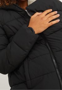 Chaqueta de abrigo negra con textura acolchada, que presenta un cuello alto y un cierre delantero con cremallera. Mano descansando suavemente sobre la tela.