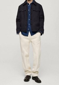 Czarna kurtka z zapięciami na guziki, niebieska koszula dżinsowa, kremowe spodnie i czarne buty. Kurtka ma kieszenie na piersi i dopasowany krój.