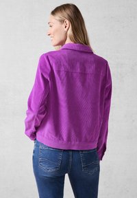 Lilla kordfløjlskjorte med lange ærmer, knaplukning ved håndleddene og krave, parret med mørkeblå jeans med baglommer og syninger.