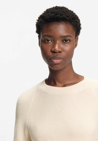 Ung svart kvinna med kort lockigt hår som bär en krämfärgad texturerad stickad tröja mot en vit bakgrund.