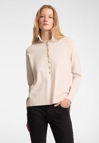 Pullover beige a maglia con colletto alto, dotato di una patta con bottoni e orli a coste, abbinato a pantaloni neri. Texture morbida e vestibilità rilassata.