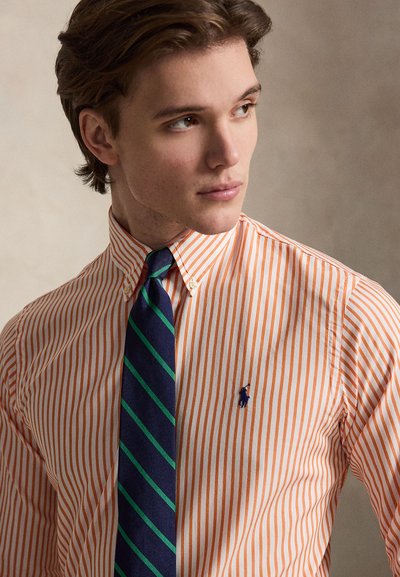 Jeune homme regardant vers la droite, portant une chemise rayée orange et blanche avec une cravate rayée bleu marine et verte.