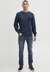 Felpa blu navy con una tasca a toppa testurizzata, polsini e orlo a costine, abbinata a jeans in denim a gamba dritta e sneakers nere.