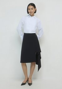 Donna con capelli corti, che indossa una camicia bianca a maniche lunghe, una gonna nera asimmetrica con dettagli drappeggiati e scarpe con tacco a punta nere, in piedi con le mani dietro la schiena.