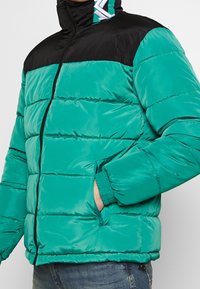 Vadderad jacka i teal med en svart övre del. Har en hög krage, dragkedjestängning och elastiska ärmslut. Slät textur med sidofickor.