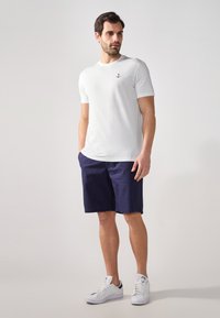 Hvid bomulds t-shirt med et sort ankerlogo, parret med marineblå shorts og hvide sneakers, stående foran en neutral baggrund.