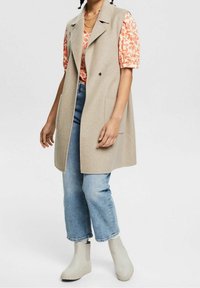 Beige Weste mit Reverskragen, ausgestattet mit zwei Vordertaschen, über einem floral gemusterten Oberteil getragen, kombiniert mit blauen Cropped-Jeans und hellgrauen Stiefeln.