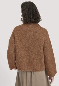 Person mit schulterlangem, dunklem, lockigem Haar, der einen lockeren braunen gestrickten Pullover und einen beigen Rock trägt, von hinten gezeigt.