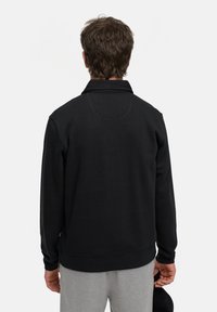 Schwarzer Fleece-Pullover mit Kragen, der ein rund gesticktes Detail auf der Rückseite und gerippte Bündchen aufweist, kombiniert mit grauen Jogginghosen.