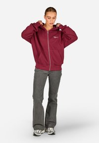 Burgundi Zip-up megztinis su raukšlėta tekstūra, priekinėmis kišenėmis ir siuvinėtu logotipu. Derinamas su pilkais platėjančiais džinsais ir sidabriniais sportbačiais.