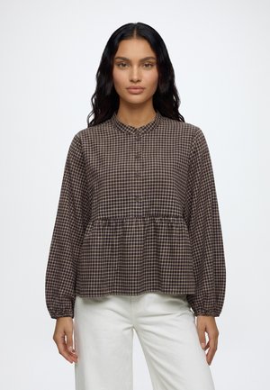 Woman with long dark hair wearing a brown checked buttoned blouse with gathered waist and white pants, standing against a plain background.