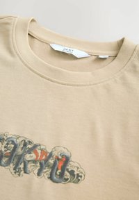 Camiseta de algodón beige con un corte relajado. Presenta un diseño bordado colorido de "Tokio" con olas y acentos en azul y rojo.