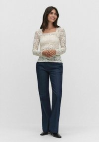 Lace långärmad topp i krämvit med fyrkantig halsringning, tillsammans med mörkblå vida byxor. Toppens design har ett texturerat spetsmönster.