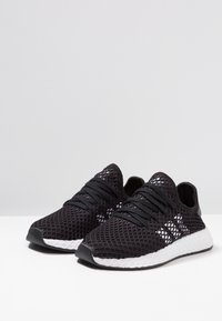 Svarta mesh-sneakers med en texturerad design, föreställande svarta snören, en svart gummisula och en vit mönstrad mellansula.