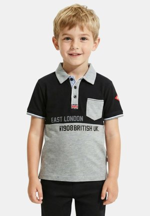 Jeune garçon blond portant un polo noir et gris avec le texte "East London 1908 British UK", debout devant un fond uni.