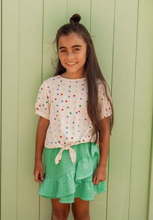 Lachend meisje met lang donker haar, gekleed in een wit shirt met kleurrijke hartjes en een gestrikte zoom, en een groene geruite rok, staand tegen een lichtgroene muur.