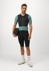 Herrcykeloutfit i svart med teal-accenter; har korta ärmar, dragkedja fram och åtsittande shorts. Bärs med en svart hjälm och vita skor.