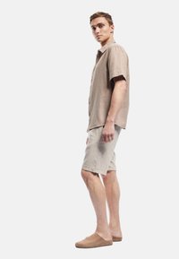 Beige kortærmet skjorte med knaplukning, parret med lyse beige shorts. Modellen har slidte tan sko på, hvilket viser en afslappet pasform.