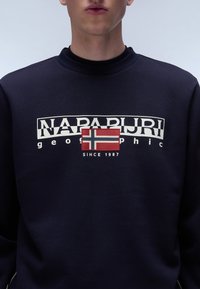 Marineblauwe sweatshirt met witte grafische tekst "NAPAPIJRI geographic" en een ontwerp van de Noorse vlag in rood en blauw. Zacht materiaal met een ronde hals.