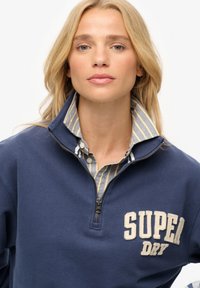 Granatowa bluza z zamkiem, z beżowym haftem "SUPER DRY", nałożona na jasnoniebieską koszulę w białe paski z guzikami.