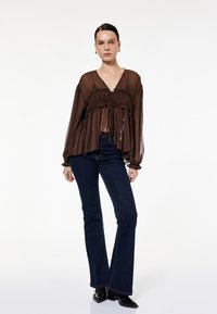 Brun, gennemsigtig bluse med lange ærmer, plisserede detaljer og V-udskæring; parret med mørkeblå flare jeans. Sorte spidse støvler.