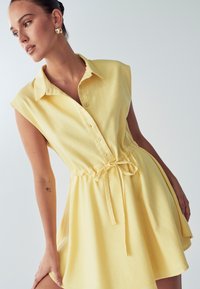 Kollane nööpidega kleit lühikeste varrukatega, lipsuga taljes ja pehme tekstuuriga. Lihtne disain, ulatub üle põlve ja flokeeritud seelikuosaga.