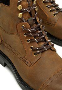 Las botas de cuero marrón presentan puntas redondas, ojales de metal y cordones con un patrón marrón y blanco. El diseño incluye costuras y un acabado texturizado.
