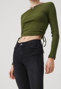 Mujer vistiendo un top recortado de manga larga arrugado en color verde oliva y unos jeans ajustados negros de cintura alta, mostrando el abdomen y un piercing en el ombligo.